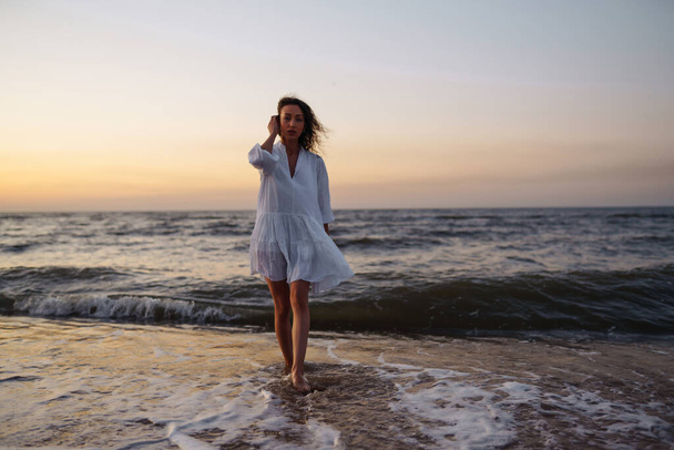 Młoda szczęśliwa kobieta w białej, trzepoczącej sukience spaceruje wzdłuż wybrzeża. Dziewczyna patrzy na magiczny wschód słońca. Czas letni. Podróże, weekend, relaks i koncepcja stylu życia. - Zdjęcie, obraz