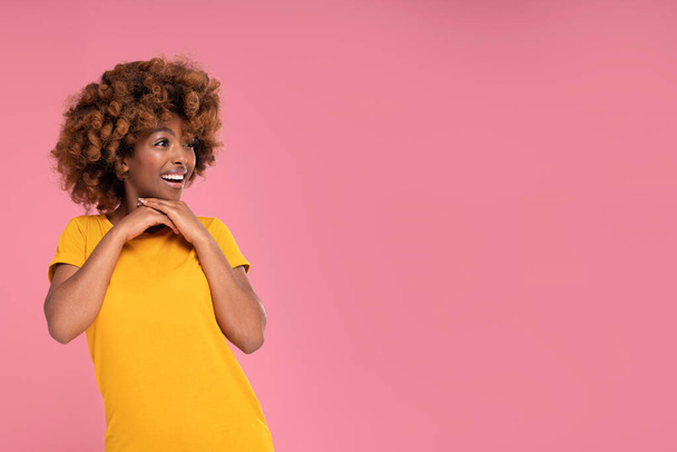 Happy beautiful afro girl smiling and looking aside on a copy space on pink pastel studio background. Colorful photo. Real people emotions. Sale. - 写真・画像
