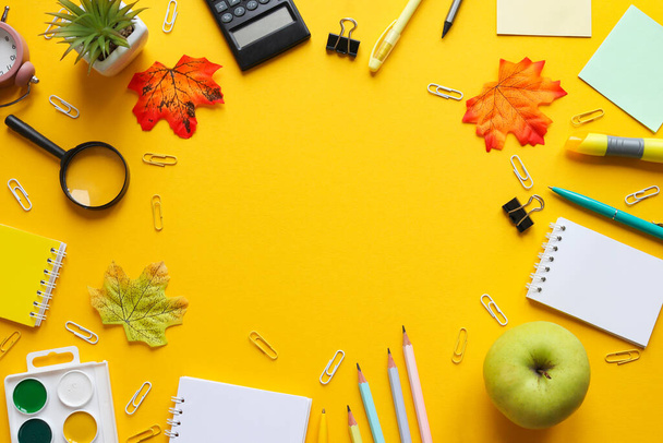 Frame of different school supplies. Notebooks, pencils and pens, paper clips, paints, a magnifying glass, and a green apple on yellow background. Copy space. Top view. Back to school concept. - Photo, Image