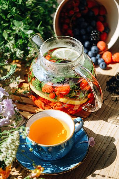 natural aromatic fruit tea in a transparent teapot and in a cup among berries and flowers. warming aromatic tea with a deep aroma of berries and wildflowers. - Foto, Imagen