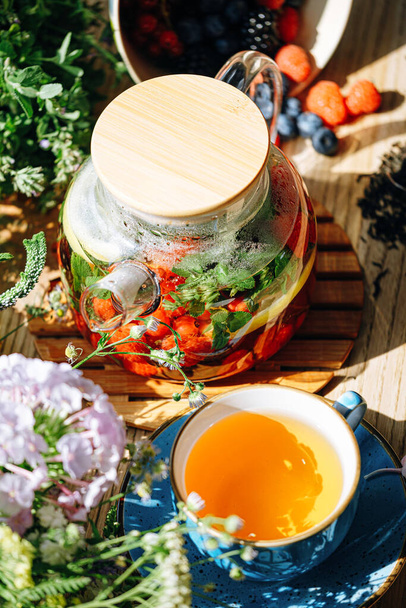 natural aromatic fruit tea in a transparent teapot and in a cup among berries and flowers. warming aromatic tea with a deep aroma of berries and wildflowers. - Fotografie, Obrázek