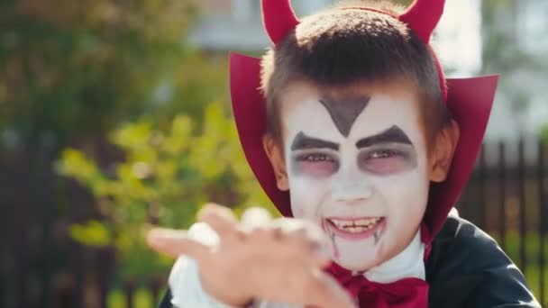 Портрет милого мальчика в макияже и костюме на Хэллоуин с дьявольскими рогами и плащом, улыбающимся и пытающимся выглядеть страшным - Кадры, видео