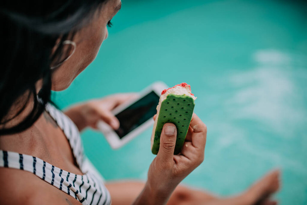 красива жінка з морозивом і смартфоном сидить біля басейну
 - Фото, зображення
