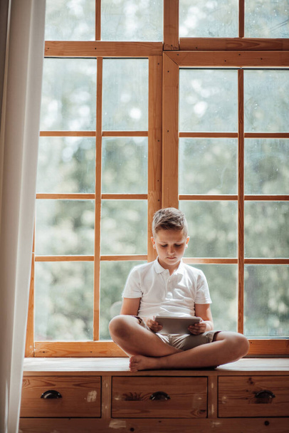 Dijital tabletli çocuk pencerenin yanında oturuyor. - Fotoğraf, Görsel