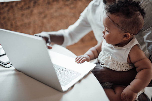 africano hombre de negocios trabajando desde casa en el ordenador portátil mientras sostiene hija - Foto, Imagen