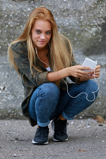 gorgeous girl listens to music in the city, portrait while looking at you with her gorgeous blue eyes, she has long blonde hair and is sporty dressed - Φωτογραφία, εικόνα