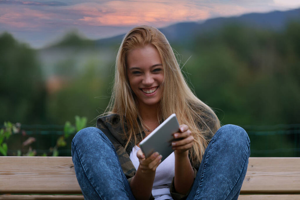 velký úsměv nebo smích, portrét dívky s tabletou na lavičce v parku při západu slunce - Fotografie, Obrázek
