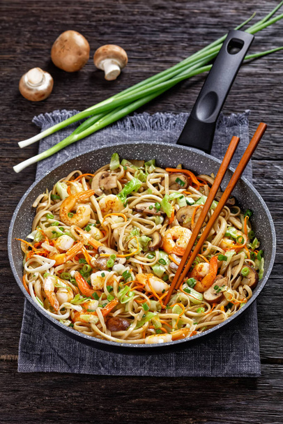 Prawn Yaki Udon, stir fried udon noodles with shrimps, cabbage, chestnut mushrooms, spring onion, carrot, mirin and soy sauce in a pan, vertical view - Foto, imagen