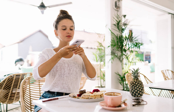 Emotionless woman in casual clothes taking photo of dish on table with cup of coffee and pineapple with smartphone in cafe - Foto, afbeelding