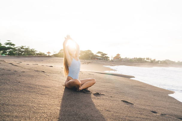 Full body anonymous female in swimwear sitting in lotus pose with raised arms on sandy beach near sea on Bali - Φωτογραφία, εικόνα