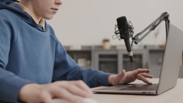 Zamknięta osoba w niebieskiej bluzie z kapturem siedząca przy biurku w domu, grająca w grę na przenośnym komputerze, rozmawiająca z profesjonalnym mikrofonem - Materiał filmowy, wideo