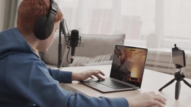 Boční pohled na rusovlasého chlapce v ušních sluchátkách sedícího za stolem v pokoji, hrající střílečku na přenosném počítači, nahrávající proces na kameře smartphonu - Záběry, video