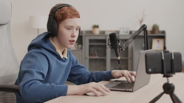 Aufmachung eines rothaarigen männlichen Influencer im Schulalter mit Kopfhörern, der am Schreibtisch in seinem Zimmer sitzt und auf einem Stativ mit einer Smartphone-Kamera spricht - Filmmaterial, Video