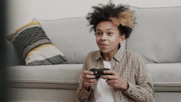 Taille-up van dertien-jarige Afrikaanse jongen zitten op de vloer door de bank thuis, met game console controller, spelen en opgewonden over het winnen - Video