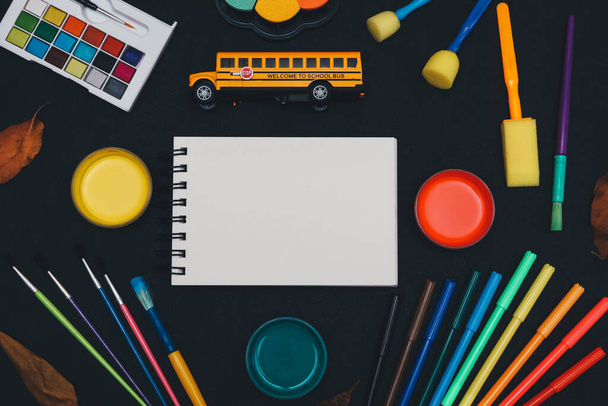 Autobús escolar, cuaderno vacío, rotuladores, pinturas y pinceles se encuentran sobre un fondo negro con espacio para el texto, vista superior de cerca. - Foto, imagen