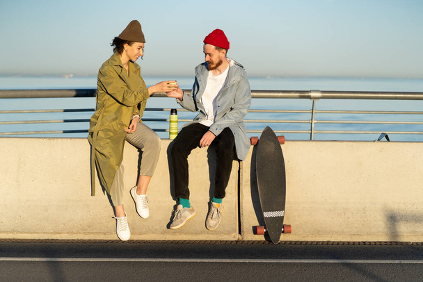 Szczęśliwa para pić gorącą herbatę na zewnątrz siedzieć na moście nosić modne ubrania ulicy z longboard w pobliżu - Zdjęcie, obraz