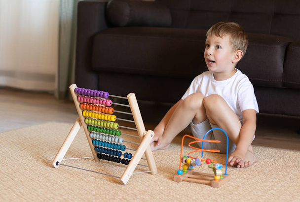 Mały chłopiec z blond włosami bawi się drewnianymi zabawkami edukacyjnymi na podłodze. Powrót do koncepcji szkoły - Zdjęcie, obraz