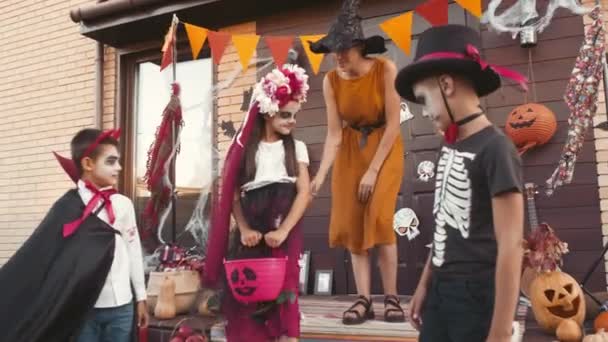 Handgehaltene Aufnahme einer jungen Frau mit Hexenhut, die auf der Veranda des Hauses steht und niedlichen Kindern in Halloween-Kostümen beim Tricksen oder Behandeln zuwinkt - Filmmaterial, Video