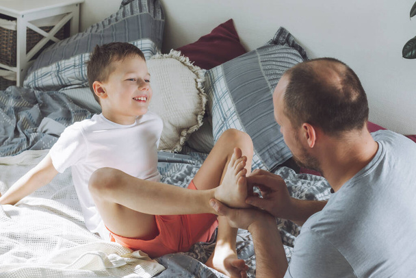 Ο πατέρας παίζει με το γιο του 7-10 στο κρεβάτι. Ο μπαμπάς γαργαλάει τα πόδια των παιδιών. Η οικογένεια διασκεδάζει. - Φωτογραφία, εικόνα