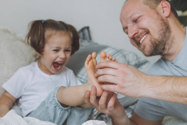 Padre juega con su hija pequeña 2-4 en la cama. Papá hace cosquillas a los niños pies. Familia, divertirse - Foto, Imagen