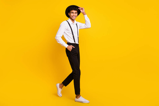 Fotografie sebevědomý chlap chůze vzhled fotoaparát nosit tričko podvazky obuv izolované žlutá barva pozadí - Fotografie, Obrázek