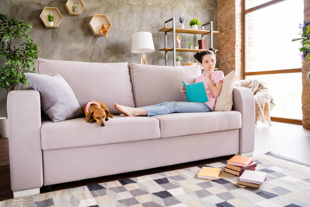 Portrait of attractive minded dreamy preteen girl reading book staying alone with doggy sitting on divan at light home house indoors - Foto, Imagem