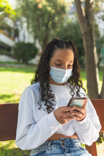 verticaal schot van een Latina tiener meisje met krullend haar dragen van een chirurgische masker met behulp van haar smartphone zittend op een park bank - Foto, afbeelding