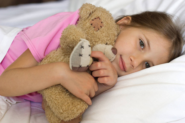 A cute little girl with blond hair in a pink T-shirt lies in bed smiling and hugging a teddy bear - Photo, Image
