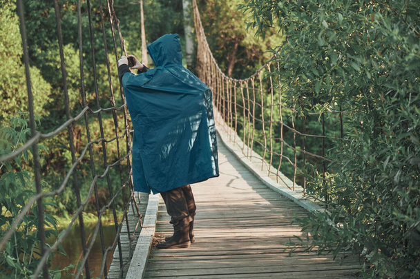 Girl hiking in raincoat on suspension bridge and takes pictures of landscape - Photo, image