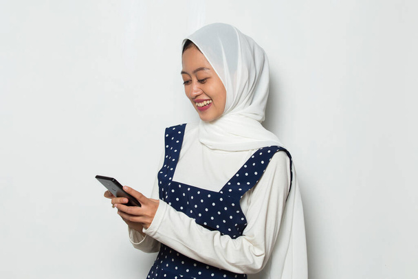счастливая молодая азиатская мусульманка с помощью мобильного телефона изолированы на белом фоне - Фото, изображение