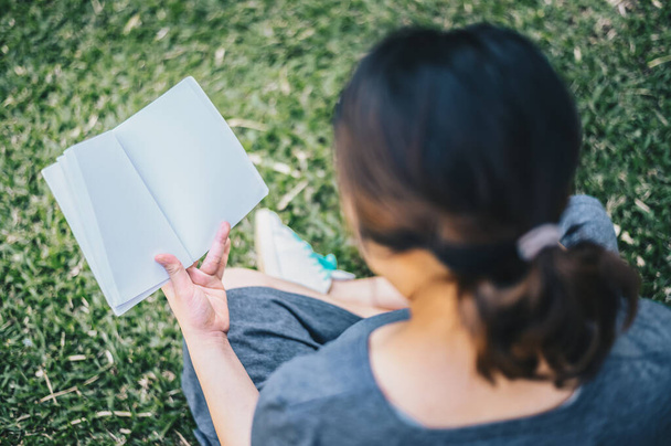 Przycięty widok na młodą ciężarną kobietę siedzącą w ogrodzie, otwartą i czytającą książkę (książka z pustą przestrzenią). Pojęcie stylu życia kobiety w ciąży. - Zdjęcie, obraz