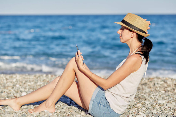  Young woman in a hat with a phone on the beach near the sea is making a selfie. Resting, traveling and relaxing concept. Copy space. High quality photo - Photo, Image