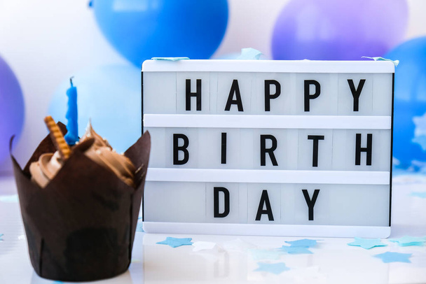 Lightbox with text HAPPY BIRTHDAY and chocolate cupcake candle on Abstract defocused blurred festive background for holiday. Blue ballons and confetti, birthday cake. Greeting card Boy concept - Photo, Image