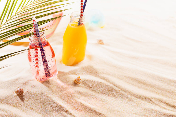 Summer tropical beach vacation background - cold fruit pink, yellow beverages in wet glass bottle with straw, hat, toy under green palm leaf on sand in sunlight, copy space. - Fotoğraf, Görsel