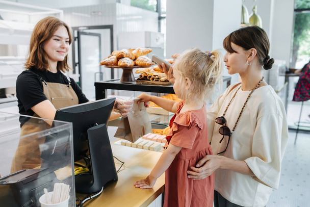 Friendly cashier handing pastry goods in a bag to mom with a daughter. In a bakery shop. Girl is standing on a step in anticipation. - Фото, изображение