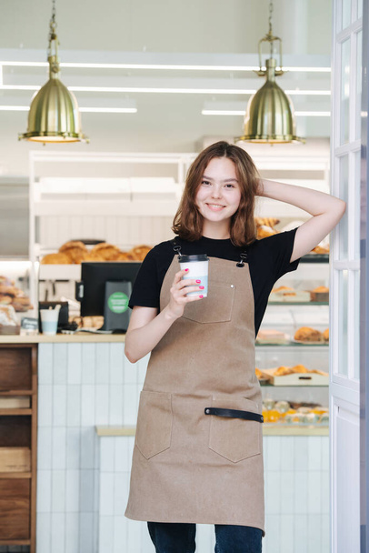 Attractive young baker girl posing with a plastic coffee cup inside her shop. She's wearing apron, looking at the camera, leaning on a door frame. Over shelves, full of pastry. - Photo, Image