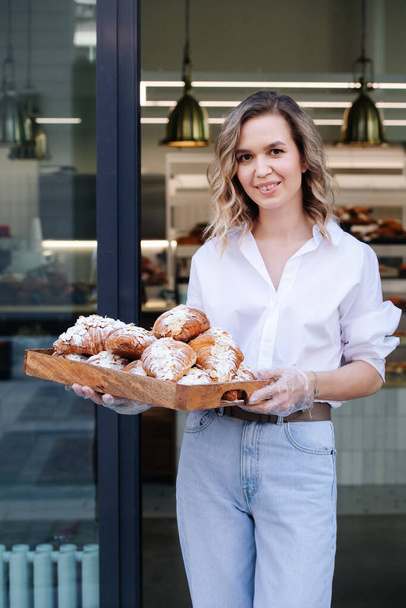 Smiling blond woman posing with a tray full of pastry in a doorstep of her shop. She's wearing dress shirt and light jeans, looking at the camera. - 写真・画像