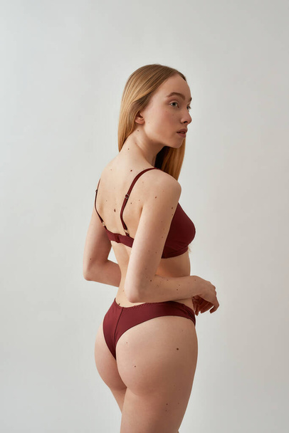 Young slim caucasian woman with beautiful fit body wearing burgundy underwear looking aside, posing isolated over light gray background - Photo, image