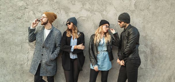 Young urban people wearing sunglasses, warm clothes and wool cap posing against a concrete wall in the street - 写真・画像