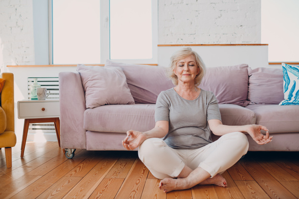 Senior adult smiling woman practicing yoga at home living room. Elderly relaxed female sitting in lotus pose and meditating zen like. - Photo, Image