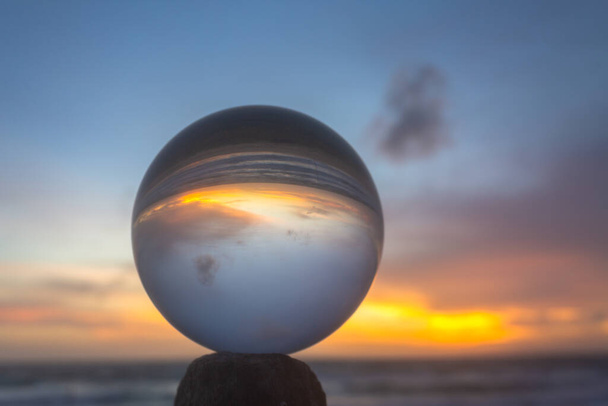 vista de la playa en el hermoso atardecer dentro de una bola de cristal colocada en una madera junto al mar. Vistas naturales poco convencionales y hermosas en la playa y puesta de sol en una bola de cristal mágico.  - Foto, imagen