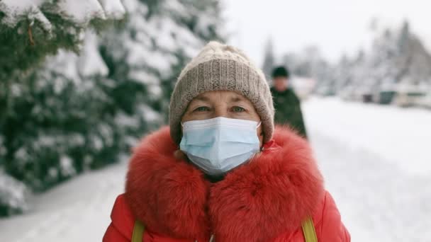 Femme âgée adulte en masque médical de protection regardant la caméra dans le parc d'hiver. Distance sociale pendant la pandémie de Coronavirus Covid 19 - Séquence, vidéo