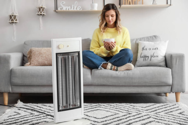 Young woman sitting in room with electric heater. Concept of heating season - Φωτογραφία, εικόνα