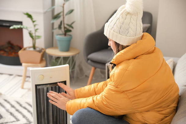 Young woman warming hands near electric heater at home. Concept of heating season - Photo, Image
