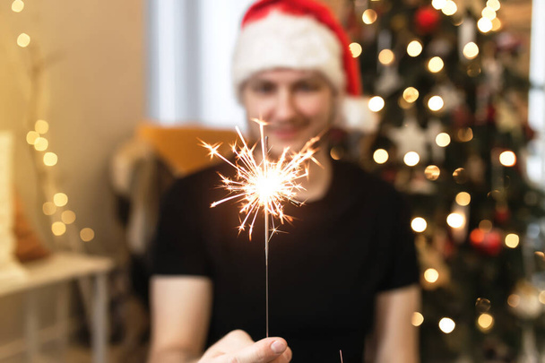 Young man in Santa hat is holding burning sparkler near Christmas tree. Christmas lights in guy hands on blurred background with decorated Christmas tree in living room. Smiling man looking at fire - Foto, Imagem
