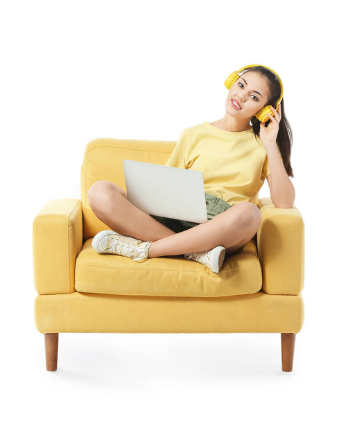 Young woman with laptop listening to music while sitting in armchair on white background - Photo, Image