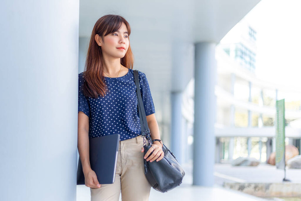 Happy young attractive asian business woman in casual clothes standing leaning against a building, holding her computer and purse while smiling at camera, young working businesswoman concept, room for copy space - Photo, Image