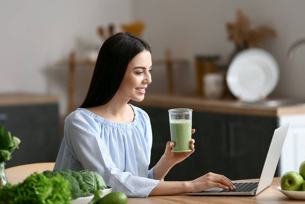 Young woman with glass of smoothie using laptop in kitchen - Фото, изображение