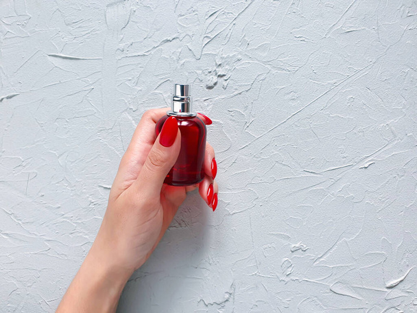 Perfume spray red bottle, female hand with red nails on gray textured background. Natural organic spa cosmetics and perfume concept. Mockup with copy space. Bottle for branding and label, front view. - Foto, afbeelding