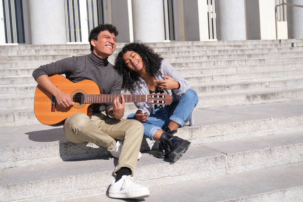 A young latin couple in love playing the guitar and singing sitting on stairs. Millennial generation. - Foto, Imagem